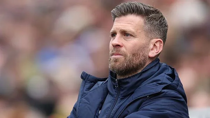 Boreham Wood manager Luke Garrard wearing a blue overcoat watching his side from the touchline at Carlisle